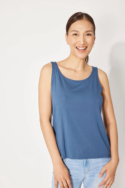 A woman wearing the Saureau Cami by Canopee in Blue, a wide-strapped tank with a scooped neck, loose fit, and hip length. She is wearing it with jeans and standing in front of a white background. 