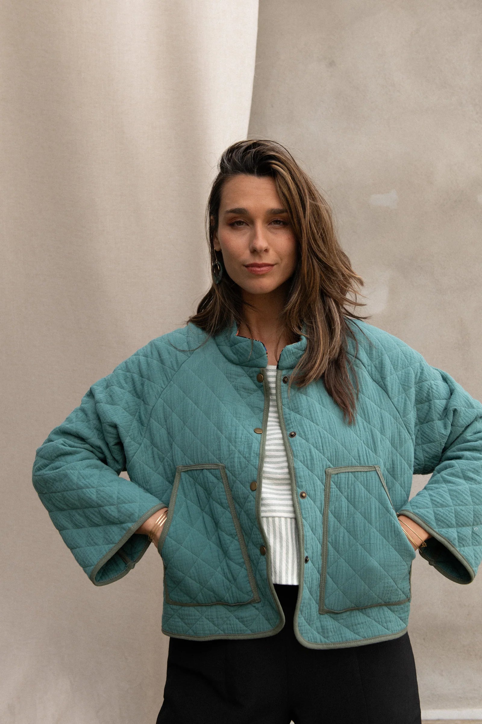 Waist up view of a woman wearing the Artemesia Jacket by Kazak in Mint, a quilted jacket with snaps up the front and large patch pockets. She is wearing it with a striped top and black pants and standing in front of a grey curtain. 