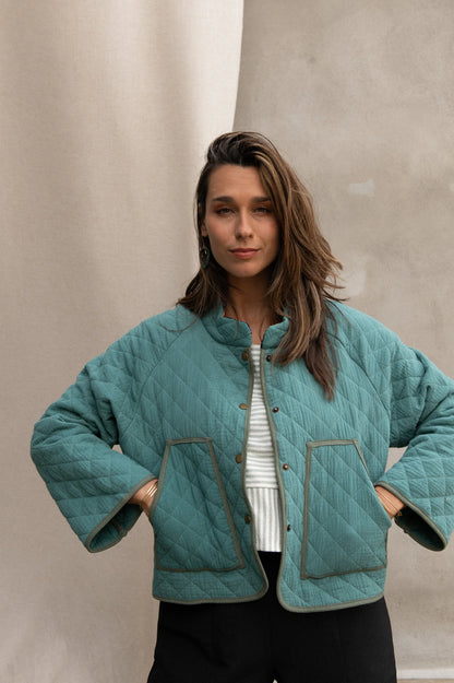 Waist up view of a woman wearing the Artemesia Jacket by Kazak in Mint, a quilted jacket with snaps up the front and large patch pockets. She is wearing it with a striped top and black pants and standing in front of a grey curtain. 