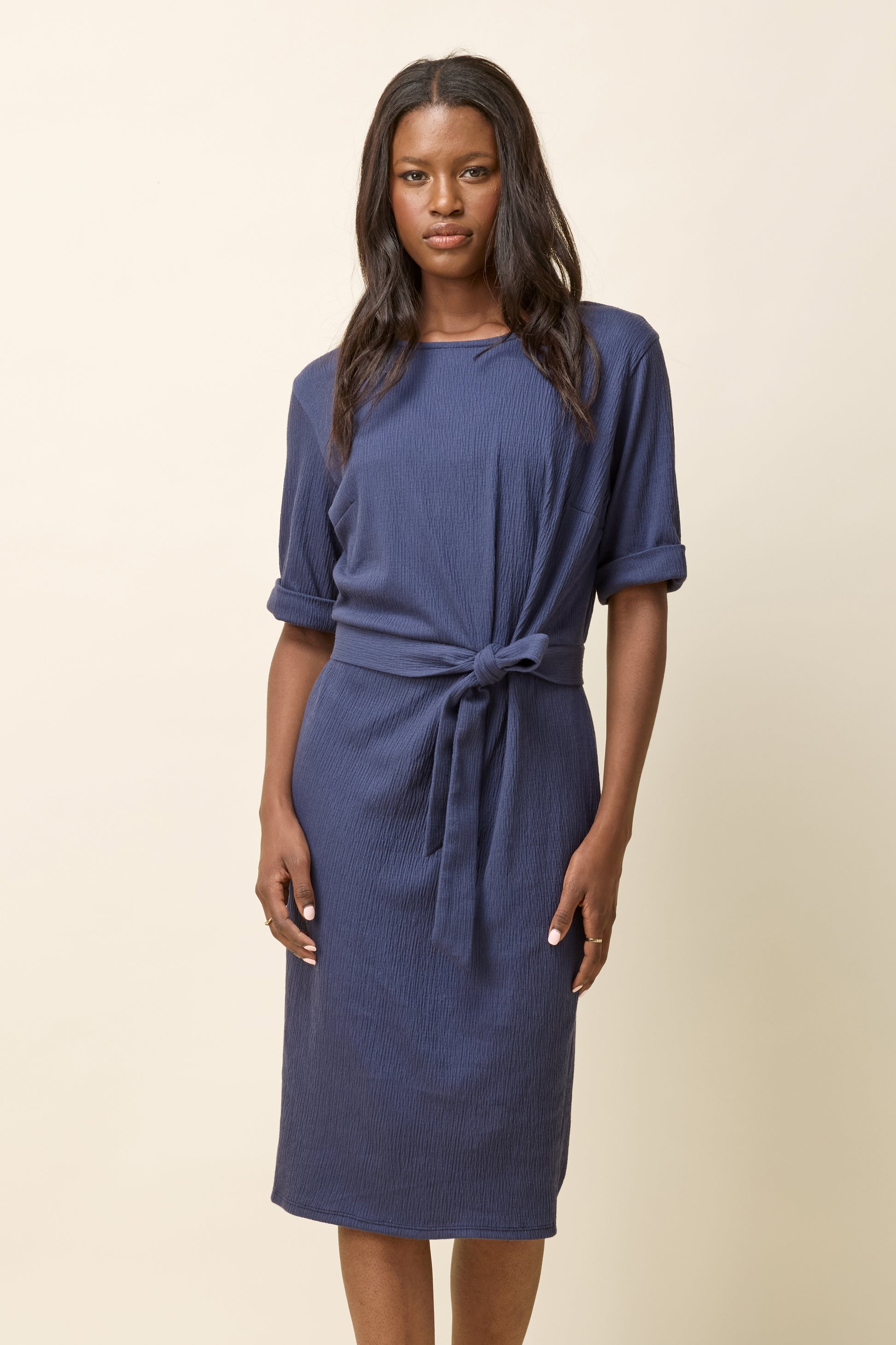 A woman wearing the Aziza Dress by Cokluch in Navy, featuring a round neck, cuffed 3/4 sleeves, an attached tie belt, and a stretchy crepe fabric. It falls below the knee. She is standing in front of a beige background. 