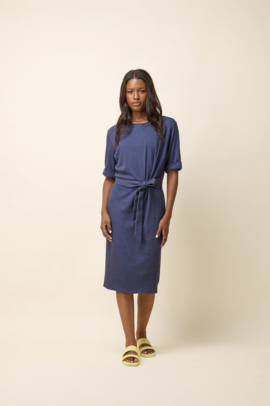 A woman wearing the Aziza Dress by Cokluch in Navy, featuring a round neck, cuffed 3/4 sleeves, an attached tie belt, and a stretchy crepe fabric. It falls below the knee. She is standing in front of a beige background. 