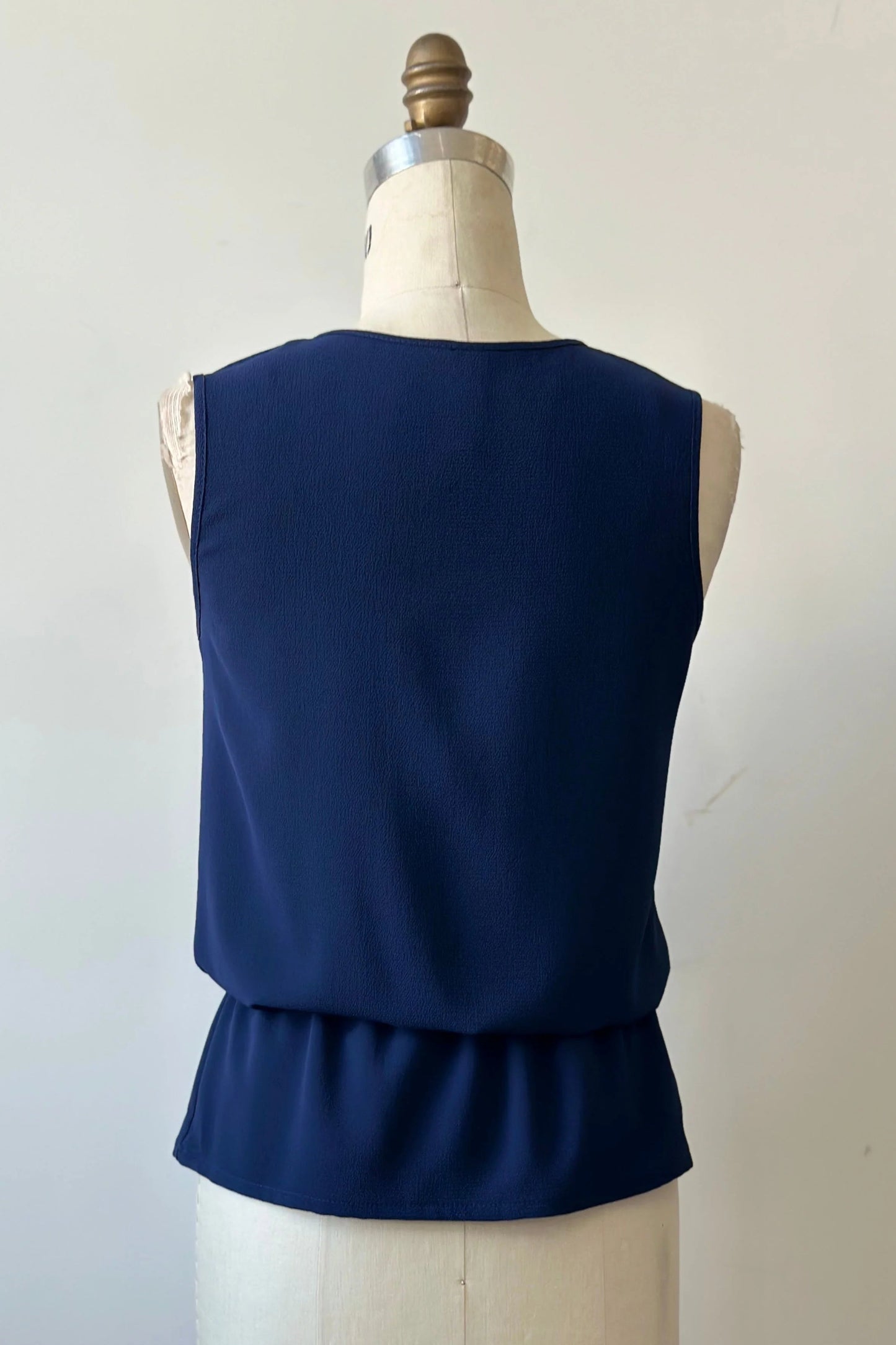 A back view of the Serena Top from Kollontai in Navy, a sleeveless top with a keyhole and tie neckline, elastic waist and flared peplum hem, is shown on a mannequin against a white background. 