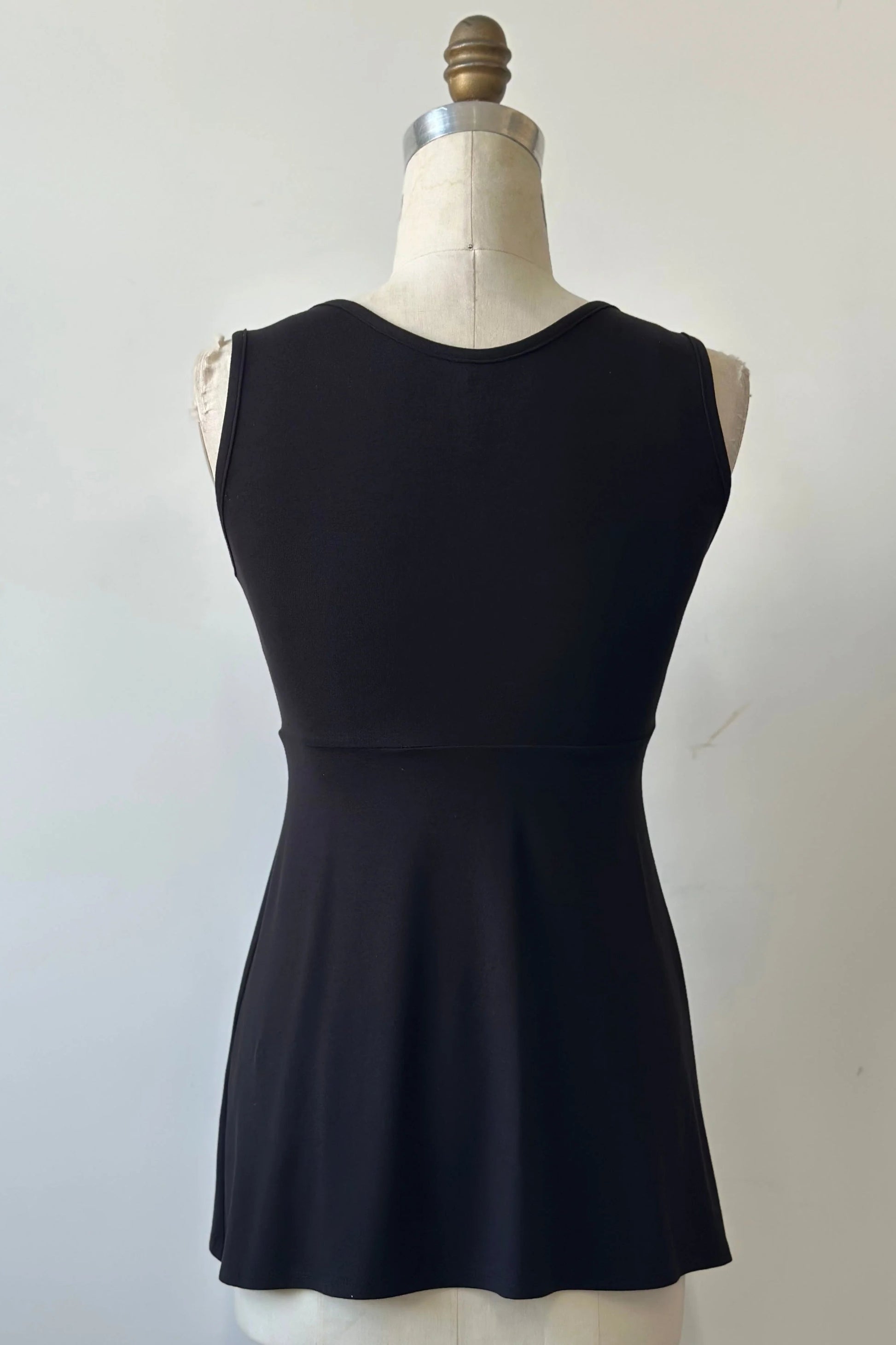 A back view of the Nira Top by Kollontai in Black, a sleeveless top with a wide neck, an empire waist, and a cut that's fitted through the bodice and flares away from the torso, is shown on a mannequin against a white background. 