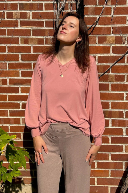 A woman wearing the Belmina Long Sleeve Top by Message Factory in Pink, with a small v-neck, long sleeves with puffed cuffs, and a relaxed fit. She is wearing it with beige pants and standing in front of a brick wall. 