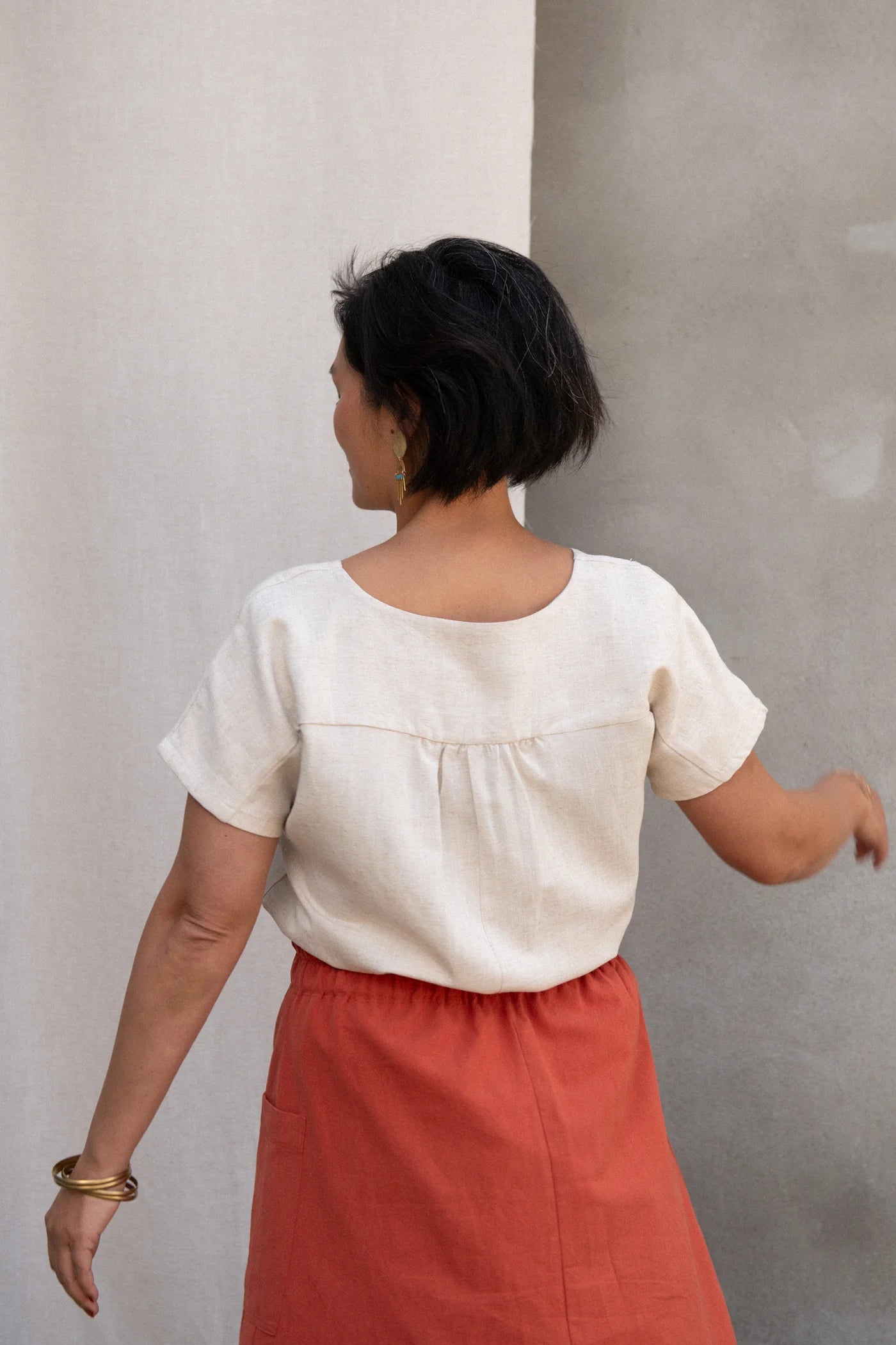 Back view of a woman wearing the Nymphea Screenprinted Skirt by Kazak in Red Pysanka with a cream top, she is standing outside in front of white curtain. 