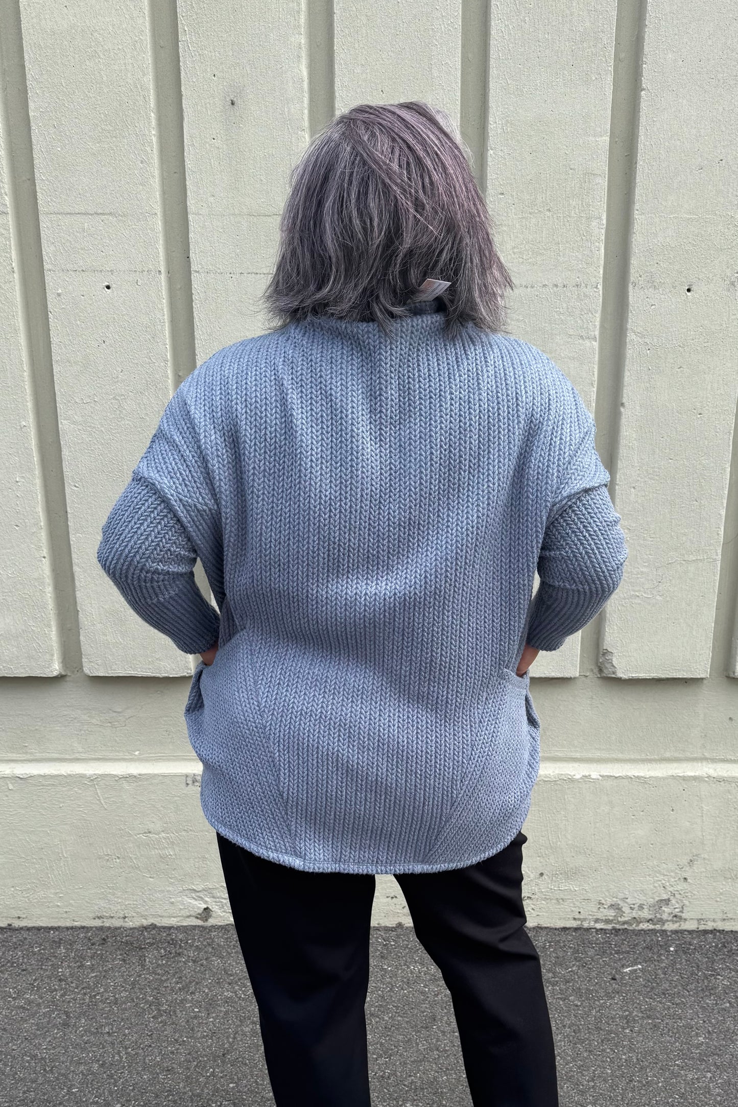Back view of a woman wearing the Meeso Sweater by MOOVment in Denim, a One Size oversized knit sweater with a wide mock turtleneck, long dropped sleeves and large side pockets. She is wearing it with black pants and standing outside in front of a grey wall. 