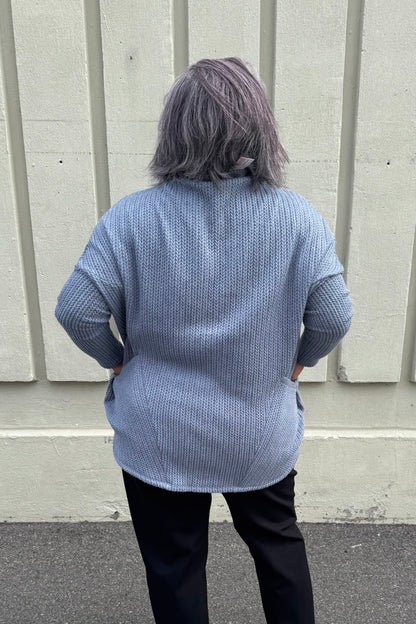 Back view of a woman wearing the Meeso Sweater by MOOVment in Denim, a One Size oversized knit sweater with a wide mock turtleneck, long dropped sleeves and large side pockets. She is wearing it with black pants and standing outside in front of a grey wall. 