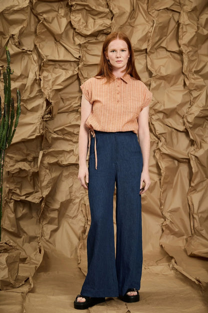 A woman wearing the Matisse Trousers by Melow, full length wide leg pants with a zipper front and a high waist with elastic at the back and side and back patch pockets. She is wearing them with a peach top and standing in front of a textured brown backdrop
