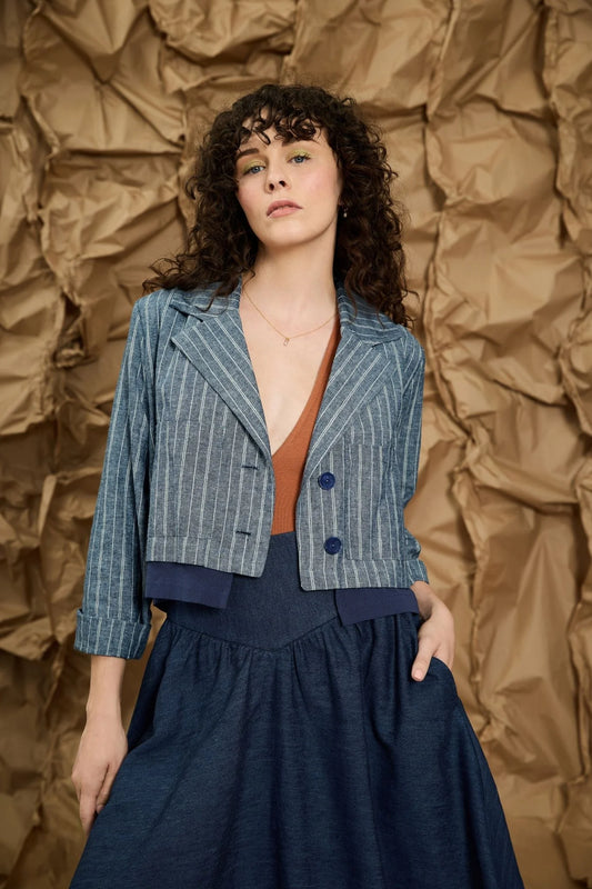 A women wearing the Maroua Jacket by Melow in Blue stripe, a cropped jacket with a tailored collar, two buttons, sleeves with a button cuff detail, and a contrasting lining band. She is wearing it with a denim skirt and an orange top and standing in front of a textured brown background. 