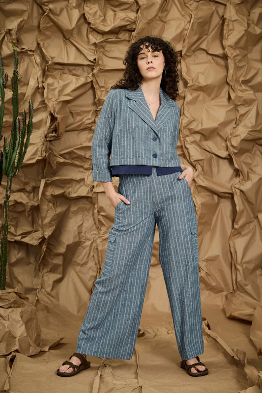 A woman wearing the Maxime Pants by Melow in Blue stripe, straight legged pants with cargo flap pockets on the legs, a high wide waistband, and an invisible side zipper. She is wearing them with a matching Marou Jacket and standing against a textured brown background