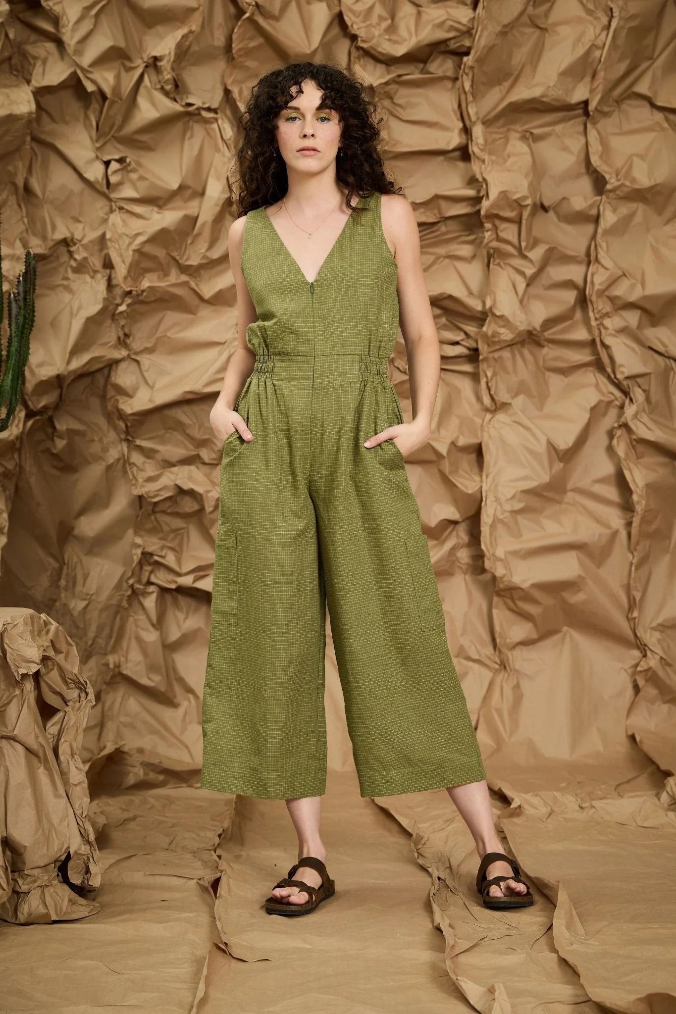 A woman wearing the Mathilde Jumpsuit by Melow in Pesto check, a sleeveless piece with a v-neck, zip front, defined waist with elastic at the sides, and wide cropped legs She is standing against a textured brown wall.