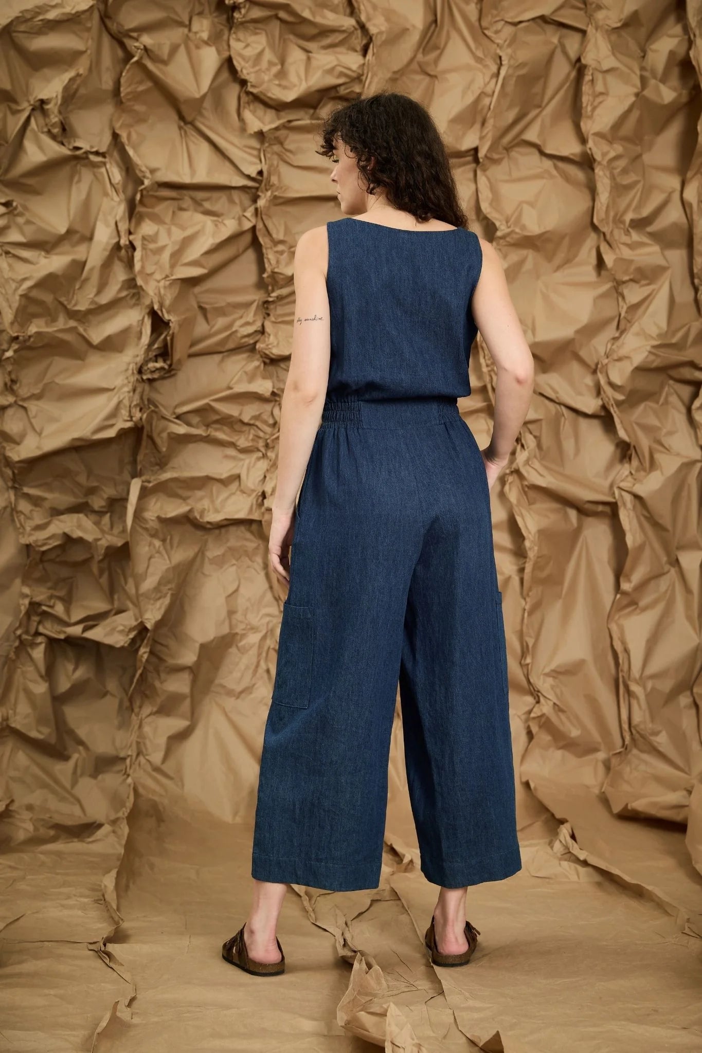 Back view of a woman wearing the Mathilde Jumpsuit by Melow in denim, a sleeveless piece with a v-neck, zip front, defined waist with elastic at the sides, and wide cropped legs She is standing against a textured brown wall.