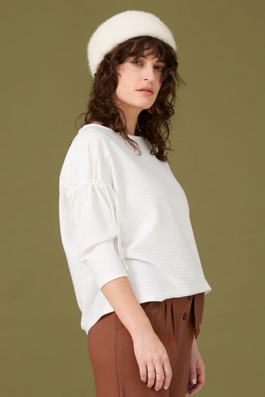  A woman wearing the Carole Top by Cokluch in Ivory, a sweater with a round neck, a drawstring detail halfway down sleeves, and a slightly loose fit. She is wearing it with brown pants and standing in front of a green background.