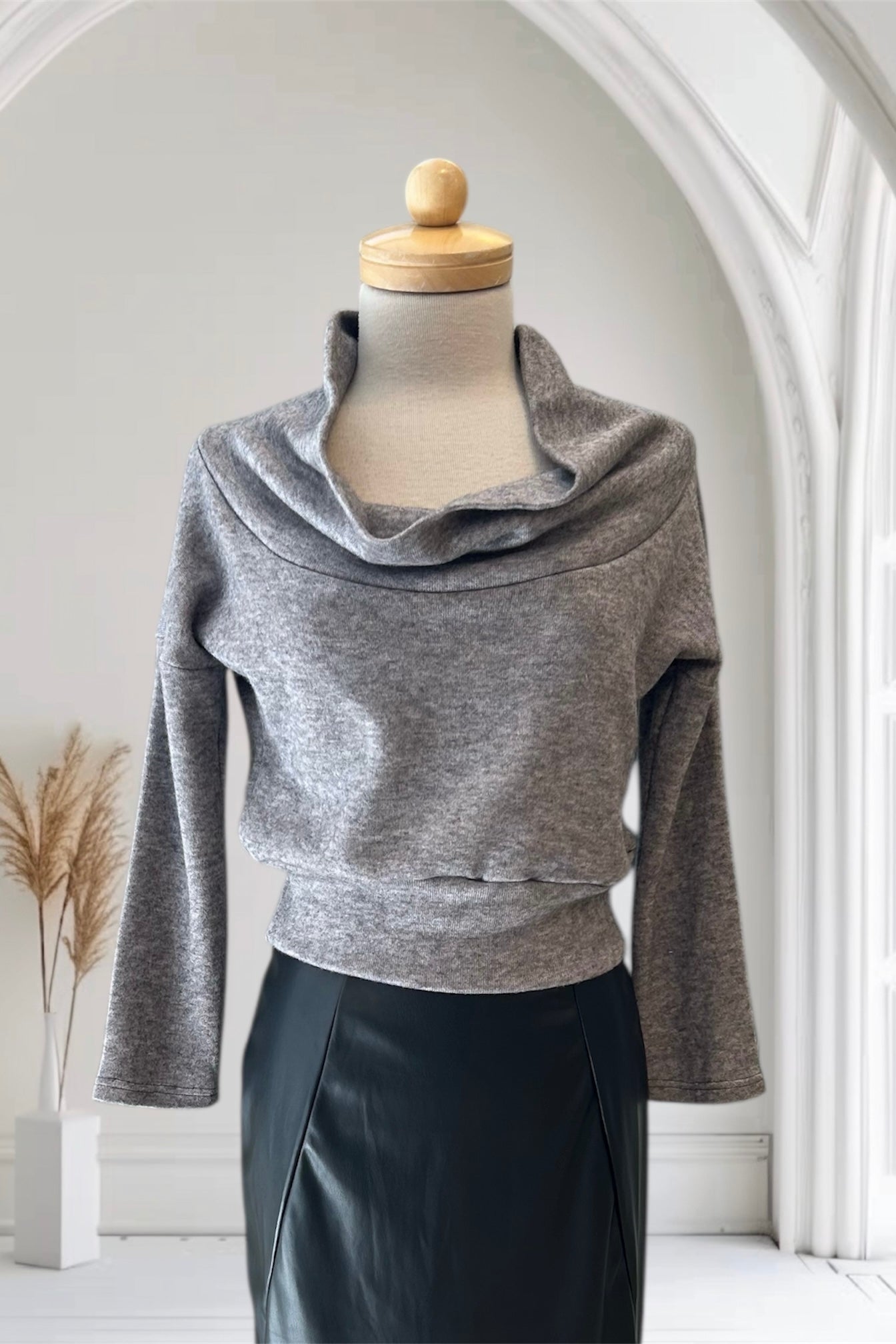 The Changes Sweater by Allison Wonderland in Light Grey, featuring a large cowl neck, long dropped sleeves, and a wide waistband, is shown with the Desire Skirt in Black against a white arched backdrop. 