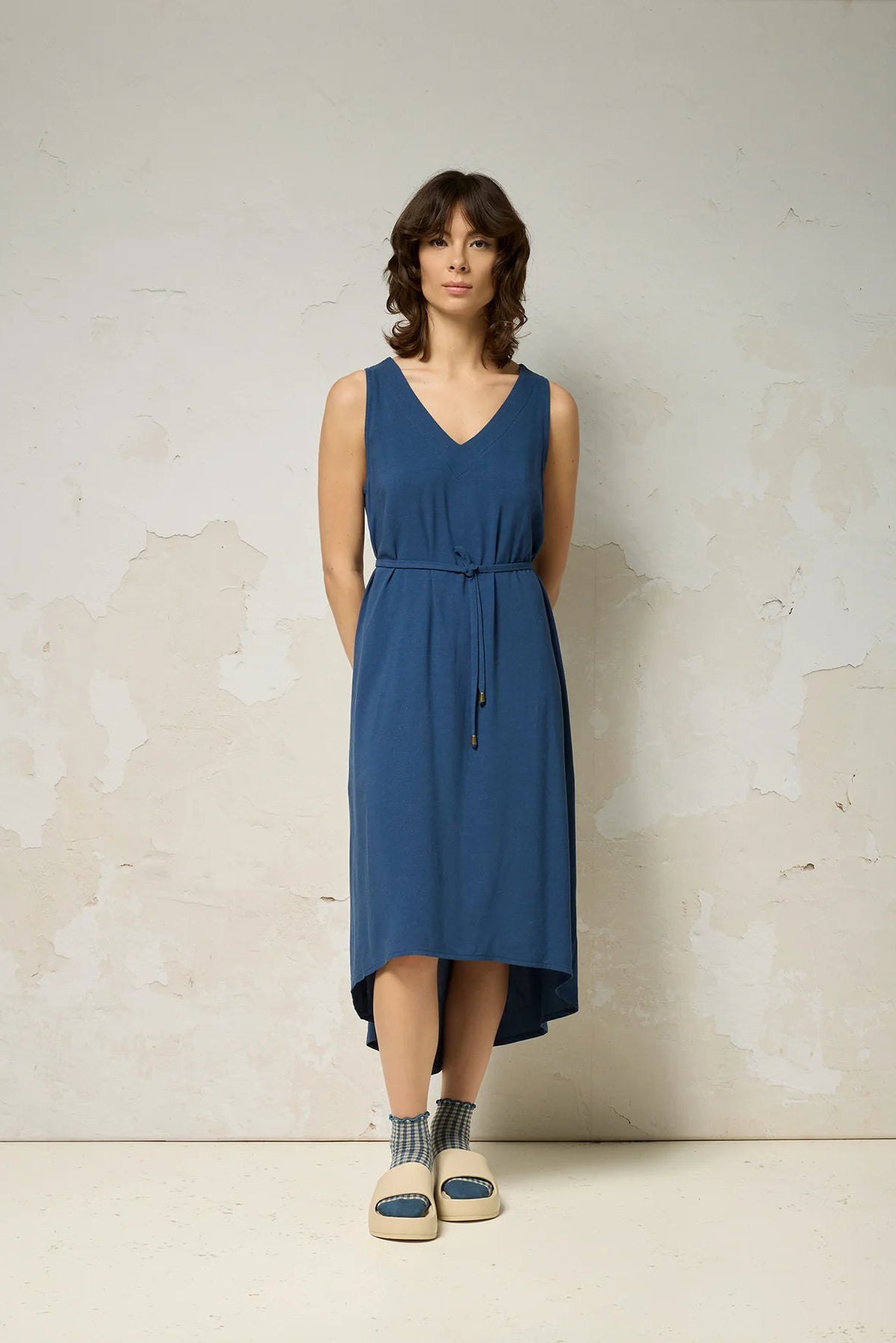 A woman wearing the Chaos Dress by Cokluch in Denim Blue, a long loose sleeveless dress with a V-neck, matching belt, and hi-low hemline. She is standing against a plain background