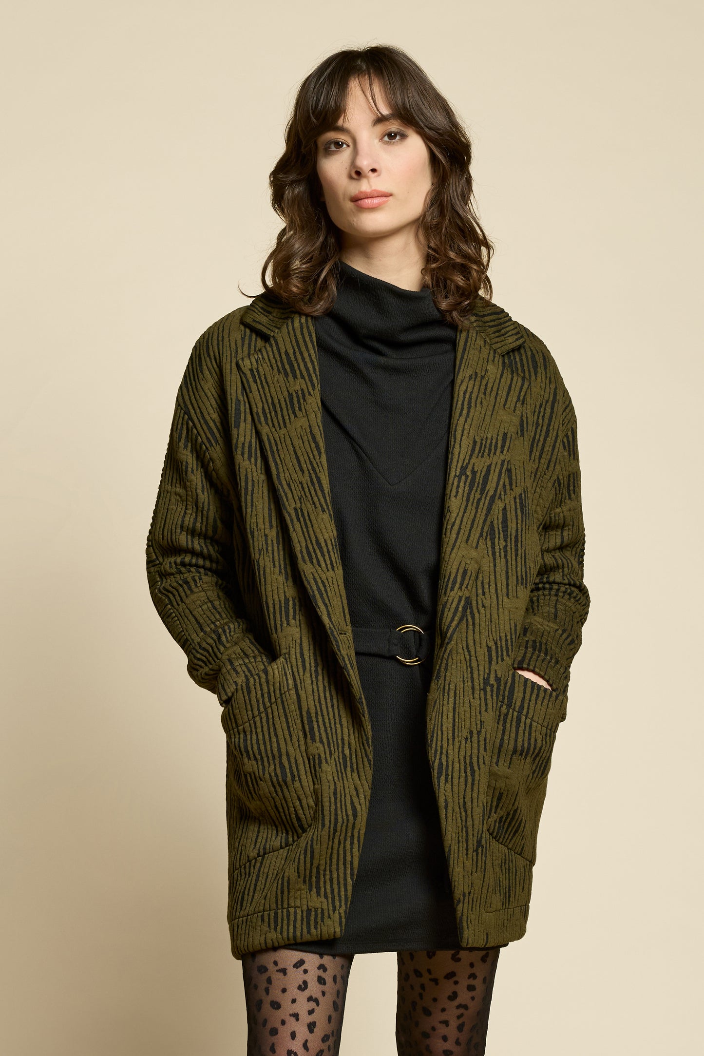 A woman wearing the Call Me Jacket by Cokluch in Olive, a thigh-length open collared jacket in unique Crinkle Max fabric with two large patch pockets. She is wearing it over a black outfit on a beige background.