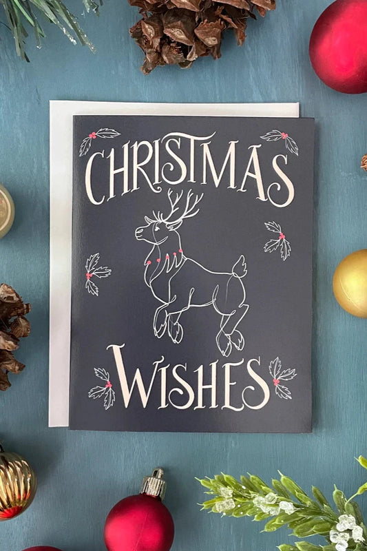 A Reindeer card with the message "Christmas Wishes" from a box set by Carabara Designs is show against a teal background with holiday decor around it. 