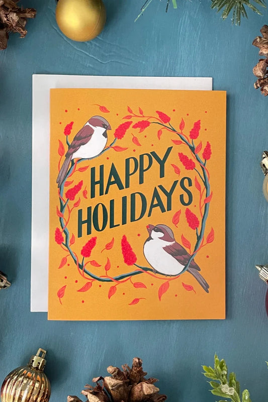 A Sparrows card from a box set by Carabara Designs, featuring sparrows and the words "Happy Holidays" on a gold background, is shown against a teal background surrounded by holiday decor. 