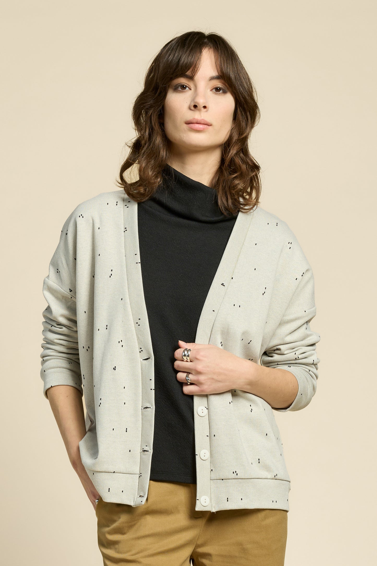Waist up view of a woman wearing the Cendres Cardigan by Cokluch in Natural, a slightly oversized cardigan in dotted fabric with four buttons. She is wearing it with a black top and yellow pants and standing in front of a beige background. 