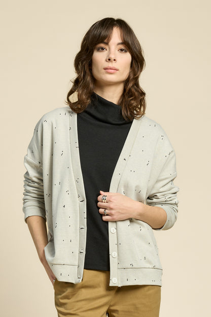 Waist up view of a woman wearing the Cendres Cardigan by Cokluch in Natural, a slightly oversized cardigan in dotted fabric with four buttons. She is wearing it with a black top and yellow pants and standing in front of a beige background. 