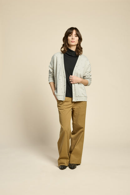 A woman wearing the Cendres Cardigan by Cokluch in Natural, a slightly oversized cardigan in dotted fabric with four buttons. She is wearing it with a black top and yellow pants and standing in front of a beige background. 