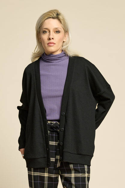 Waist up view of a woman wearing the Cendres Cardigan by Cokluch in Black, a slightly oversized cardigan in dotted fabric with four buttons. She is wearing it with a purple top and the West Coast pants in Black Plaid and standing in front of a beige background. 