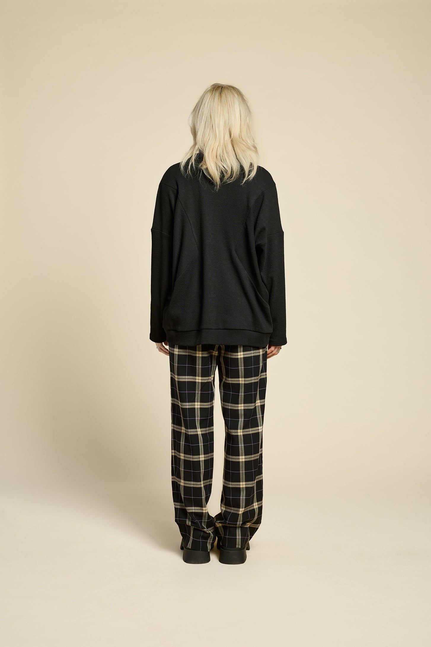 Back view of a woman wearing the Cendres Cardigan by Cokluch in Black, a slightly oversized cardigan in dotted fabric with four buttons. She is wearing it with a purple top and the West Coast pants in Black Plaid and standing in front of a beige background. 