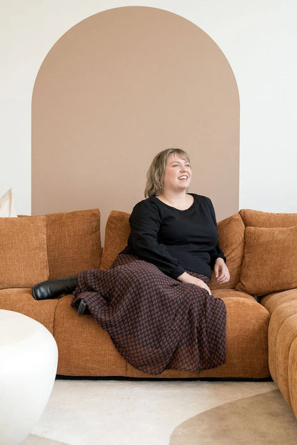 A woman wearing the Espresso Skirt by Cherry Bobin in Black and Brown print, a long and flowing chiffon skirt that's fully lined. She's wearing it with a black top and sitting on a brown sofa. 