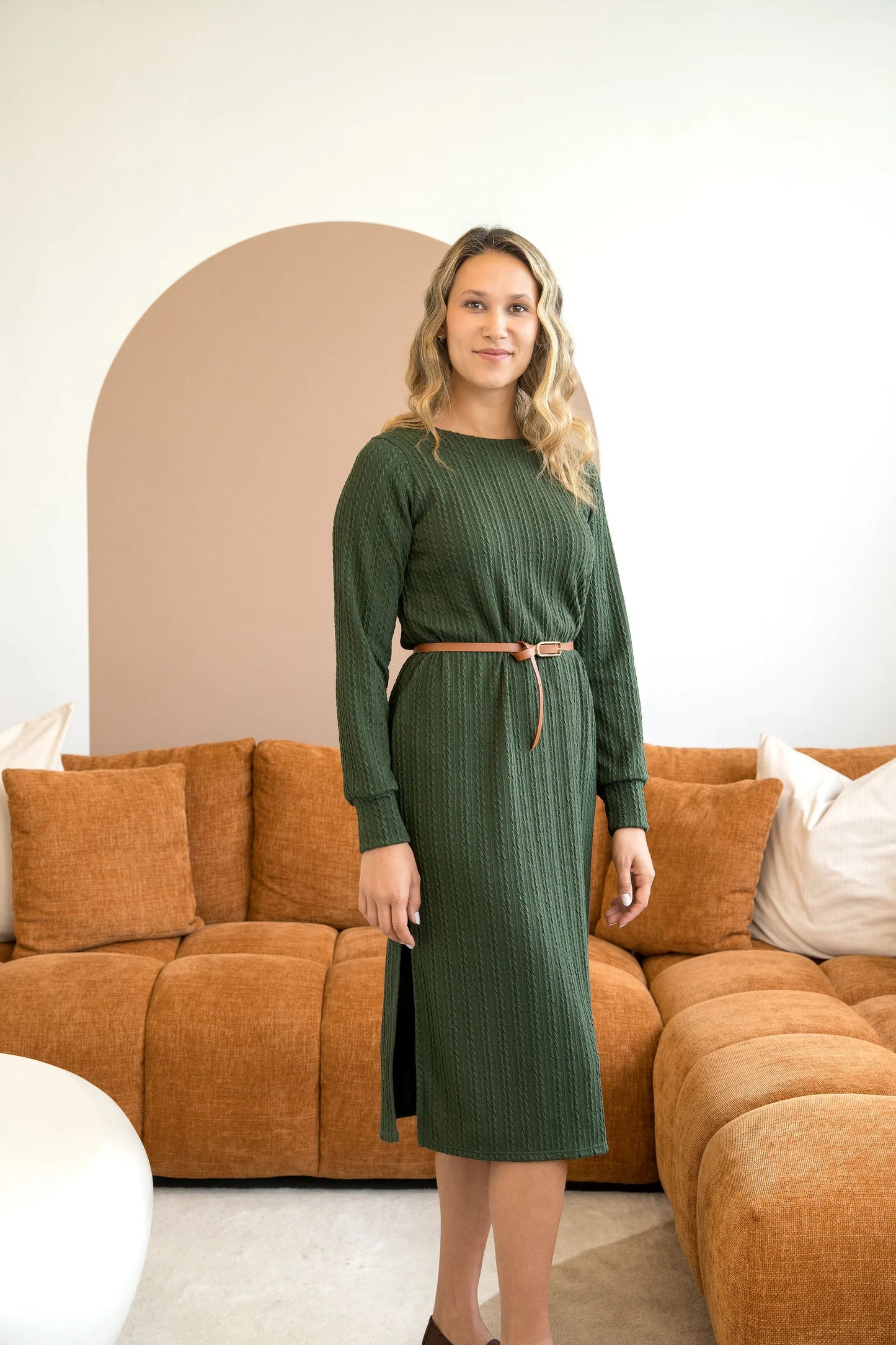 A woman wearing the Negroni Dress by Cherry Bobin in Green, a mid-calf dress with in cable-knit textured fabric, side slits, and long, slighty puffed sleeves. She is standing in a room with a brown couch. 