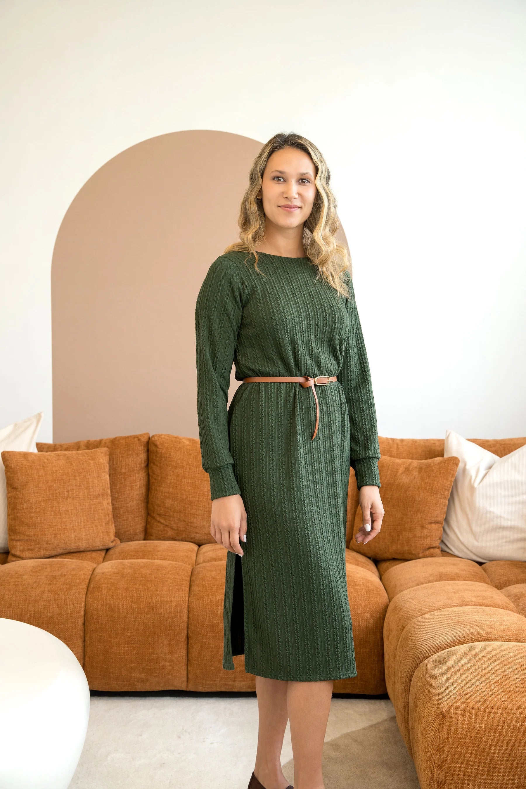 A woman wearing the Negroni Dress by Cherry Bobin in Green, a mid-calf dress with in cable-knit textured fabric, side slits, and long, slighty puffed sleeves. She is standing in a room with a brown couch. 