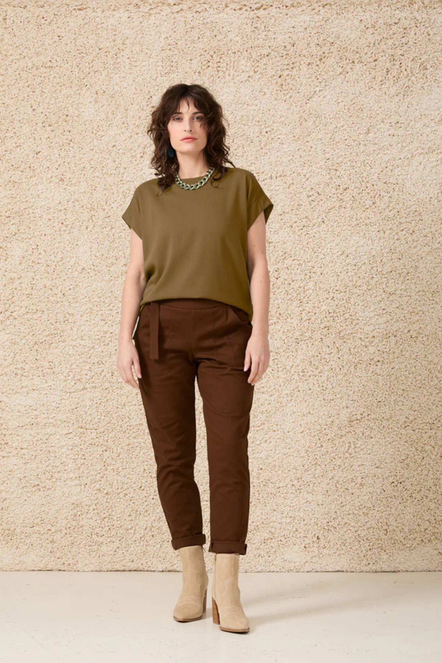 A woman wearing the Ombre Top by Cokluch in Oregano, featuring a round neck, short dropped sleeves and a loose fit, with brown pants,  standing in front of a beige wall 