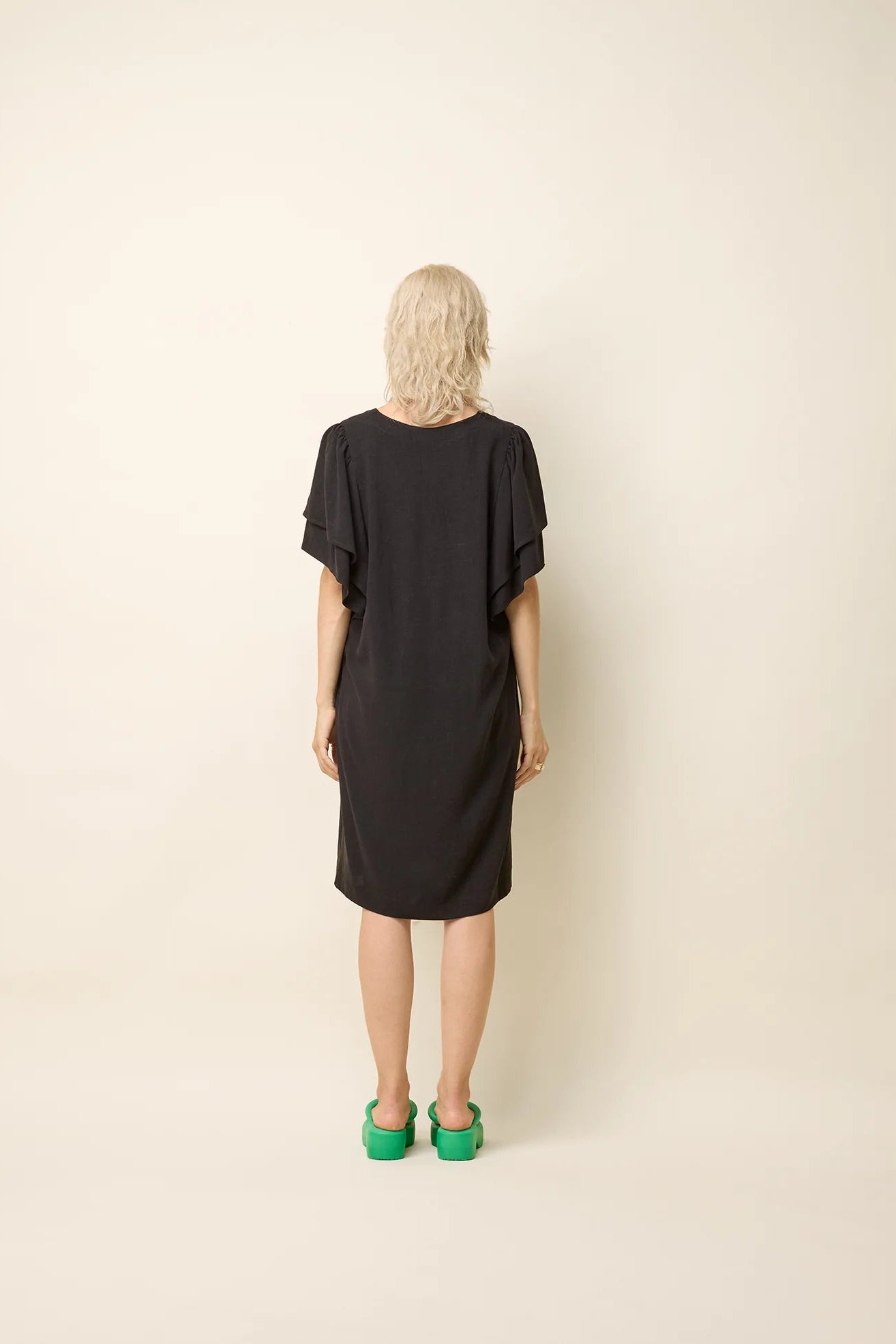 Back view of a woman wearing the Paramida Dress by Cokluch in Black, featuring a v-neck, a double ruffle on the short sleeves, and a loose fit that falls above the knee. She is standing in front of a beige background. 