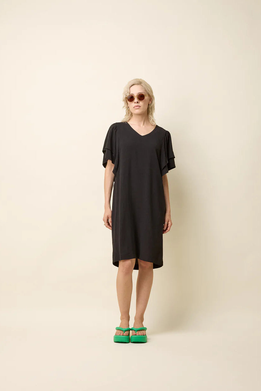 A woman wearing the Paramida Dress by Cokluch in Black, featuring a v-neck, a double ruffle on the short sleeves, and a loose fit that falls above the knee. She is standing in front of a beige background. 