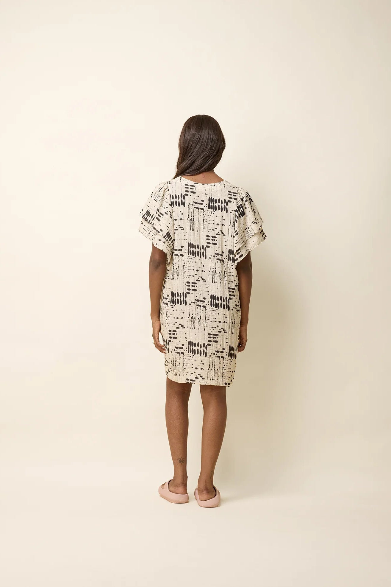 A woman wearing the Paramida Dress by Cokluch in Natural Ink, featuring a v-neck, a double ruffle on the short sleeves, and a loose fit that falls above the knee. She is standing in front of a beige background. 