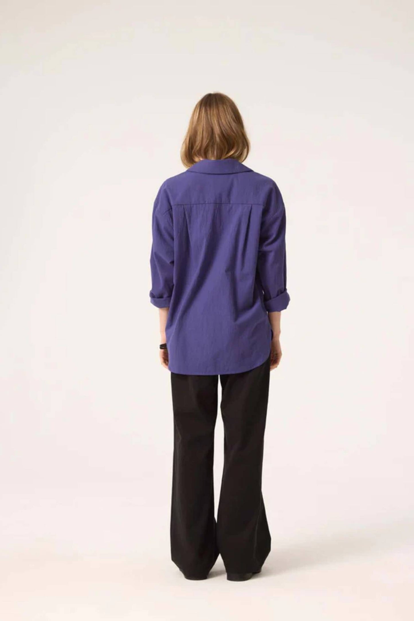 Back view of a woman wearing the Willow Top by Cokluch in Twilight with dark pants, standing in front of a white background 