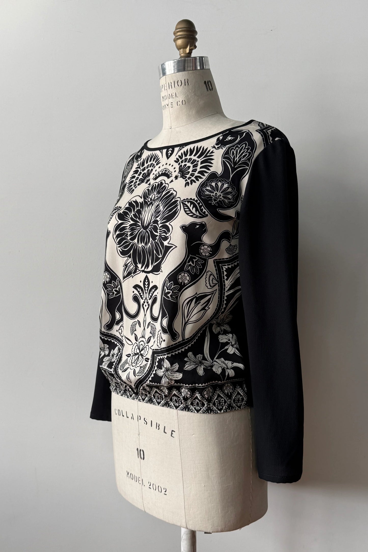 The Oriane Blouse by Kollontai, featuring a black-on-ivory floral and panther print, solid black long sleeves and back, and an elasticized hem, is shown on a mannequin against a grey background. 