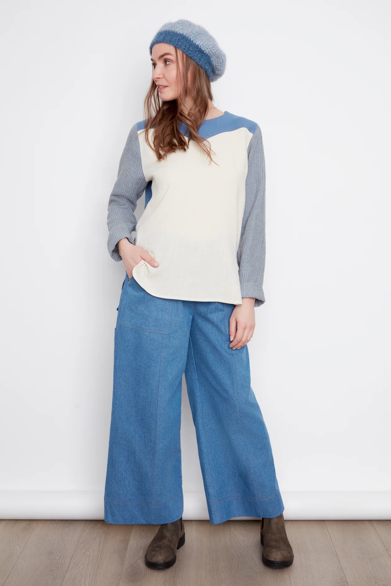 A woman wearing the Brooke Wide Leg Denim Pants by Julei in light denimm, with large patch pockets, a zipper, elastic at the back waist, and a carpenter loop. She is wearing them with a white and grey top and standing in front of a white background. 