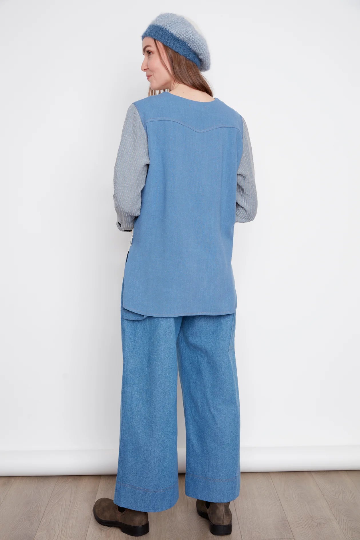 Back view of a woman wearing the Brooke Wide Leg Denim Pants by Julei in light denim, with large patch pockets, a zipper, elastic at the back waist, and a carpenter loop. She is wearing them with a white and blue and grey top and standing in front of a white background. 