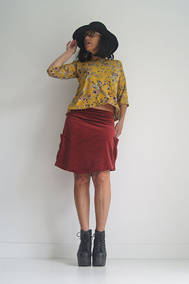 A woman wearing the Oversized Top by SI Design in Yellow Floral, featuring a scoop neck, elbow-length sleeves, and a slightly relaxed fit. She is wearing with a burgundy skirt and standing in front of a grey background. 