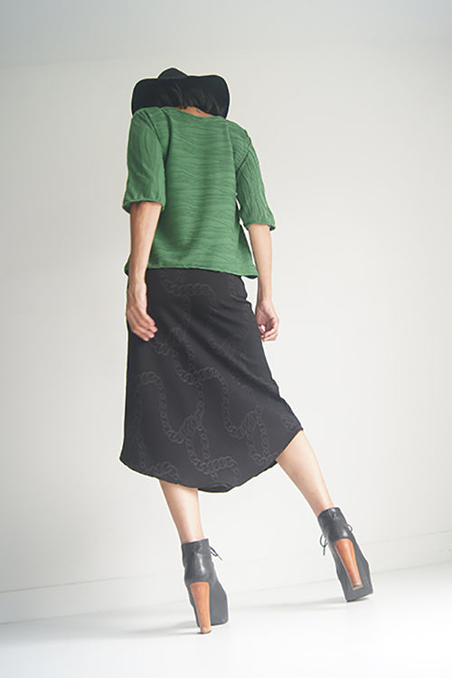 Back view of a woman wearing the Oversized Top by SI Design in Green, featuring a scoop neck, elbow-length sleeves, and a slightly relaxed fit. She is wearing with a black skirt and standing in front of a grey background. 