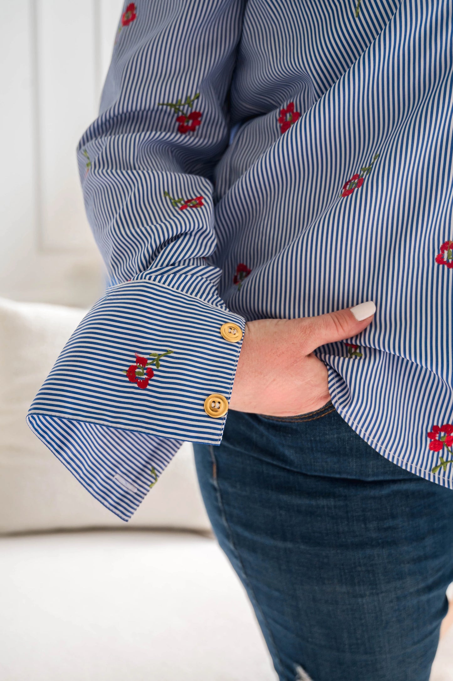 Close up sleeve and fabric detail of a woman wearing the Stripes and Roses shirt by Dotty in navy/white stripe with embroidered red roses, a collar, vintage gold buttons up the front, and double button cuffs. She is wearing it with jeans and standing in front of a white sofa. 