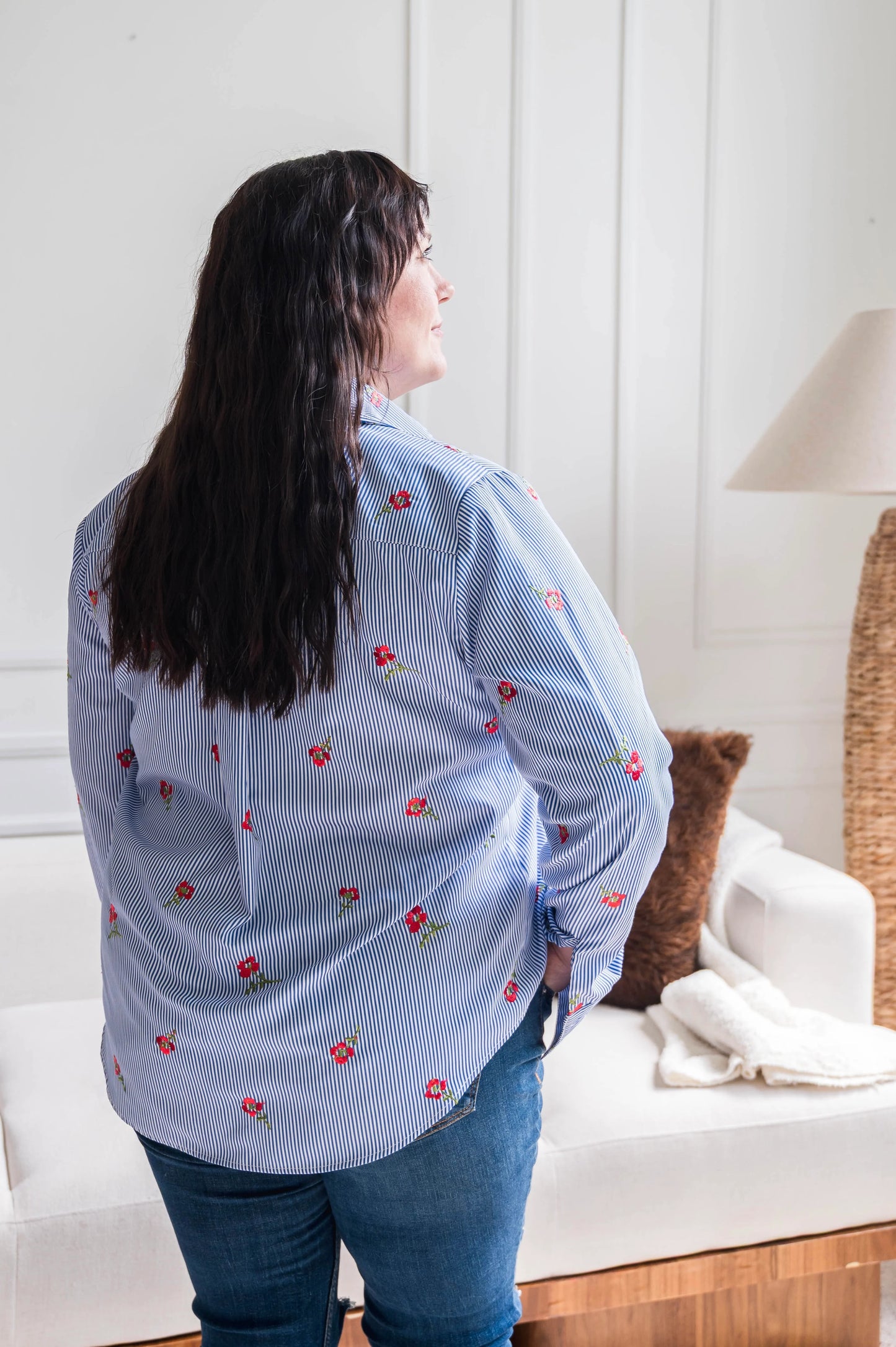 Back view of a woman wearing the Stripes and Roses shirt by Dotty in navy/white stripe with embroidered red roses, a collar, vintage gold buttons up the front, and double button cuffs. She is wearing it with jeans and standing in front of a white sofa. 