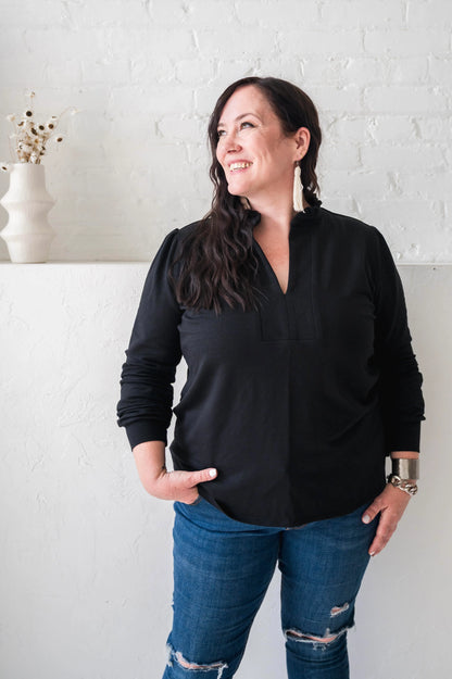 A woman wearing the Sweatshirt Blouse by Dotty in Black, featuring a ruffled v-neck, long sleeves with banded cuffs, and a relaxed fit. She is wearing it with jeans and standing in front of a white wall. 