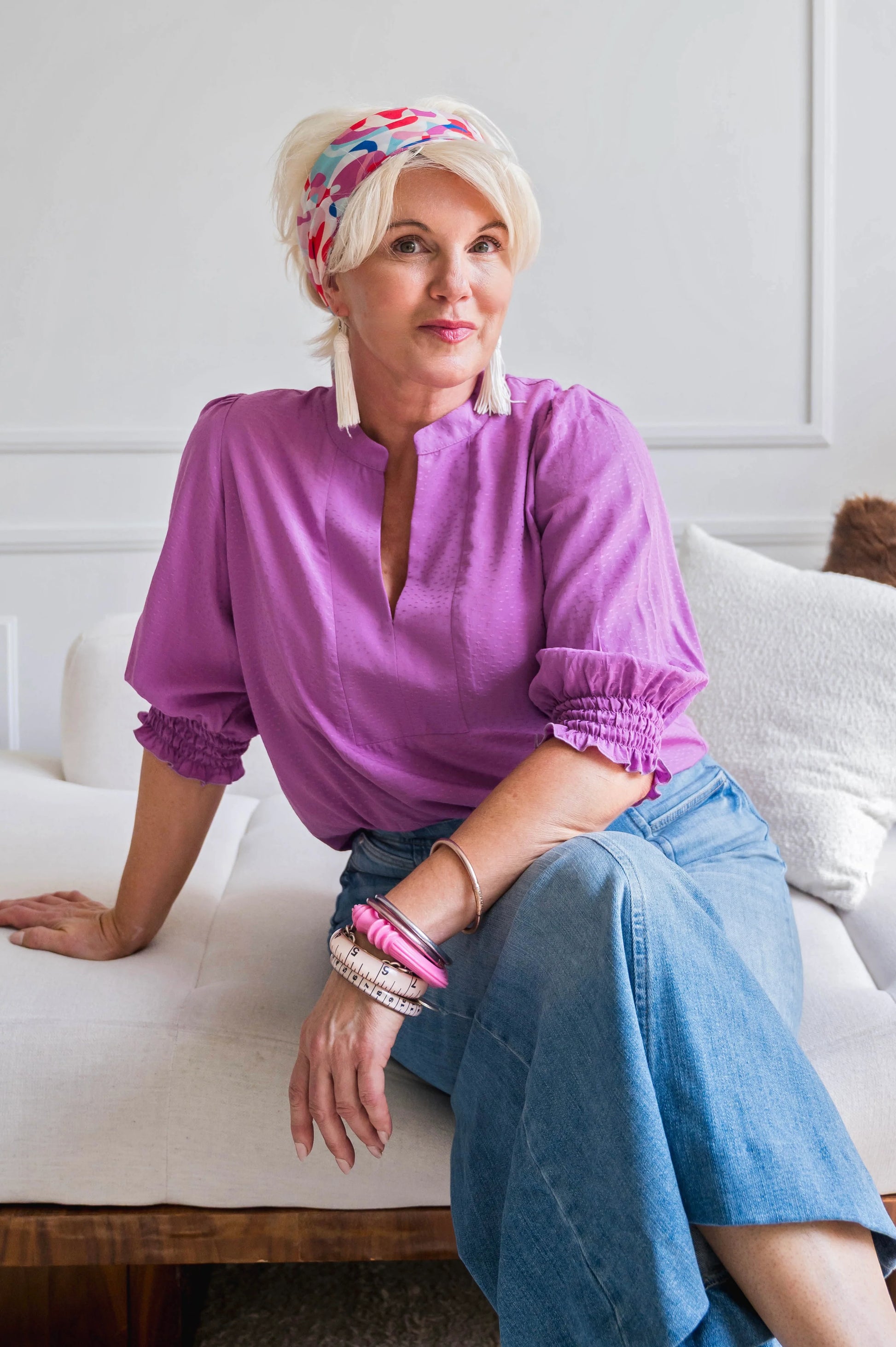 A woman wearing the Orchid Blouse by Dotty in Magenta, with a V-neck with a banded collar and elbow length smocked sleeves. She is wearing it with jeans and sitting on a couch.