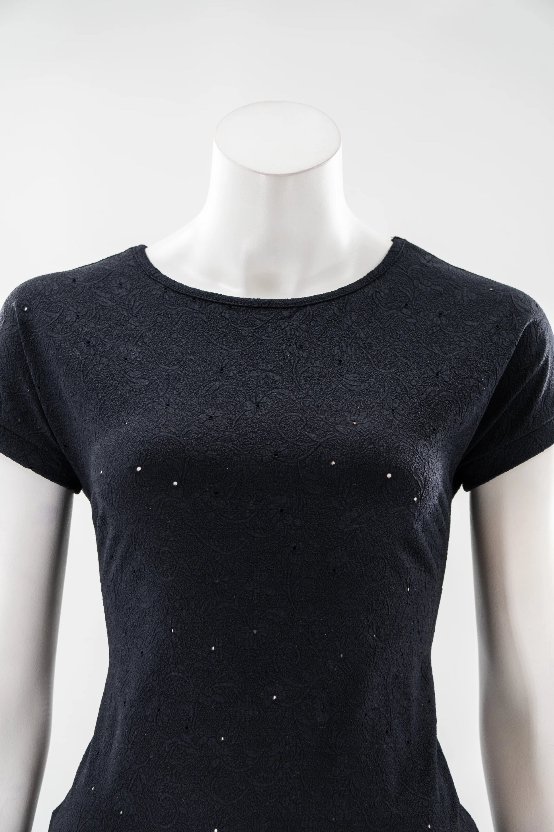 The Tourmaline T-Shirt by Karkass in Black, featuring a subtle floral texture with tiny cut-outs, is show on a mannequin in front of a white background. 