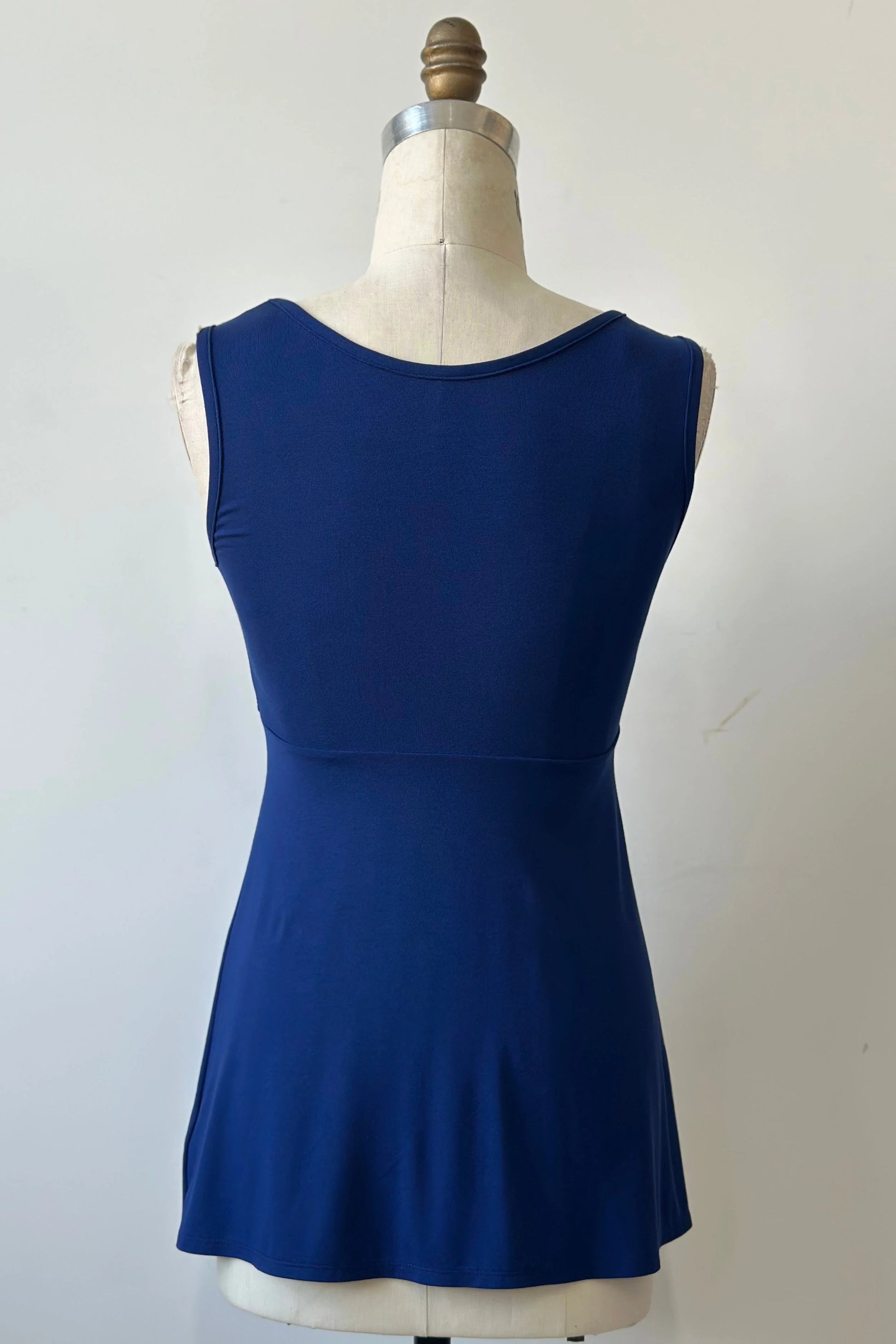 A back view of the Nira Top by Kollontai in Admiral blue, a sleeveless top with a wide neck, an empire waist, and a cut that's fitted through the bodice and flares away from the torso, is shown on a mannequin against a white background. 