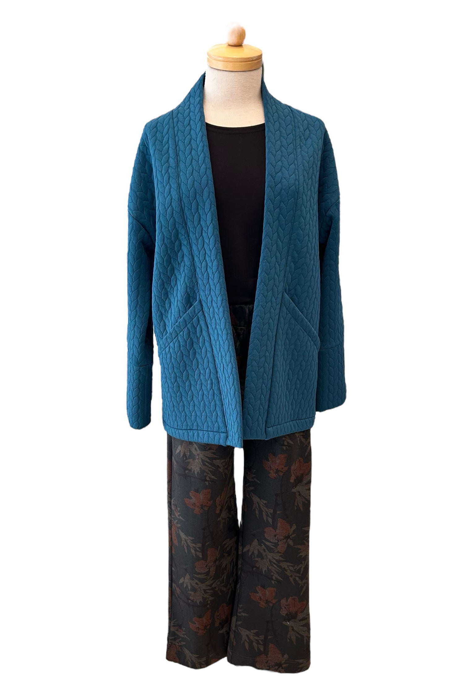 The Ina Coatigan by Tangente in Peacock, an open front cardigan in a cable-knit textured fabric, with dropped sleeves and patch pockets, is shown on a mannequin with patterned pants against a white background. 