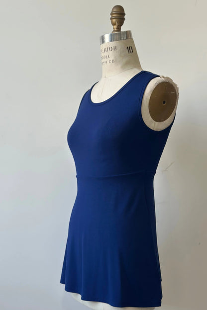 A side view of the Nira Top by Kollontai in Admiral blue, a sleeveless top with a wide neck, an empire waist, and a cut that's fitted through the bodice and flares away from the torso, is shown on a mannequin against a white background. 