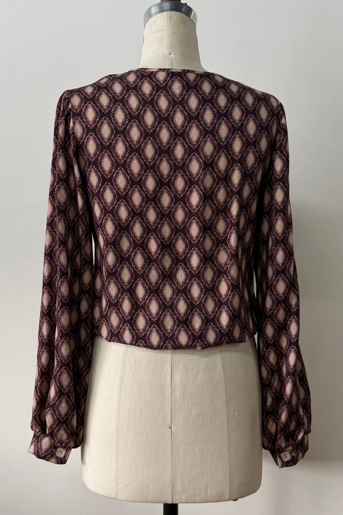 A back view of the Isabella Blouse by Kollontai, a burgundy and pink patterned blouse with a tie detail at the neck, balloon sleeves with square jewel buttons, and an elastic waist, is shown on a mannequin against a white background. 