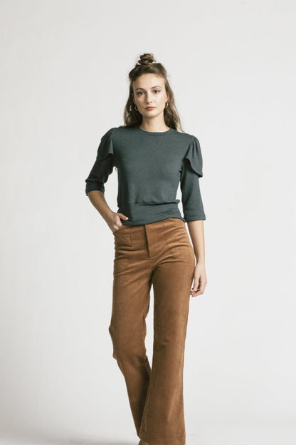 A woman wearing the Eyre Sweater by Allison Wonderland in Denim, with brown pants, standing in front of a white background 